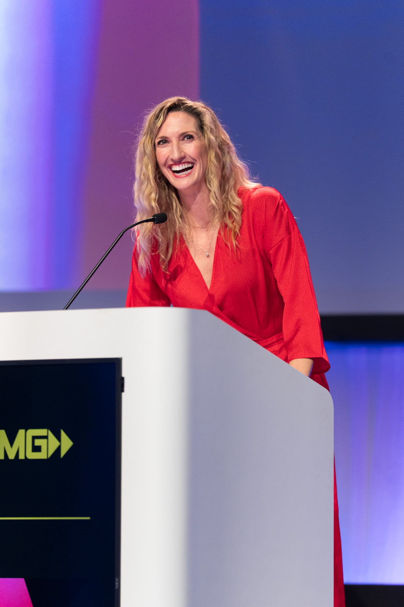 A host in a red dress laughing at the lectern at the Film G Awards