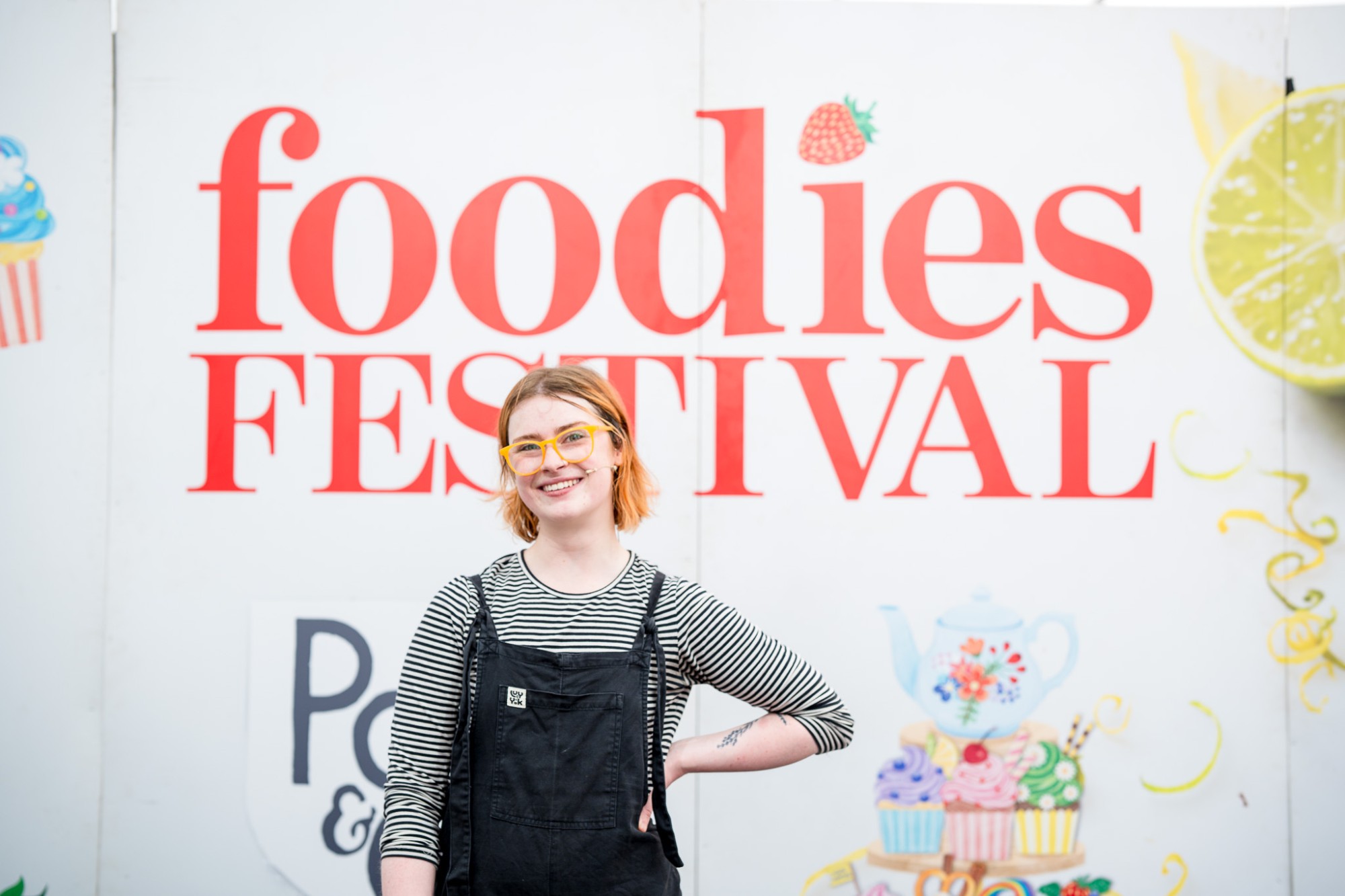 A smiling woman in yellow glasses and dungarees in front of the Foodies Festival sign in Glasgow