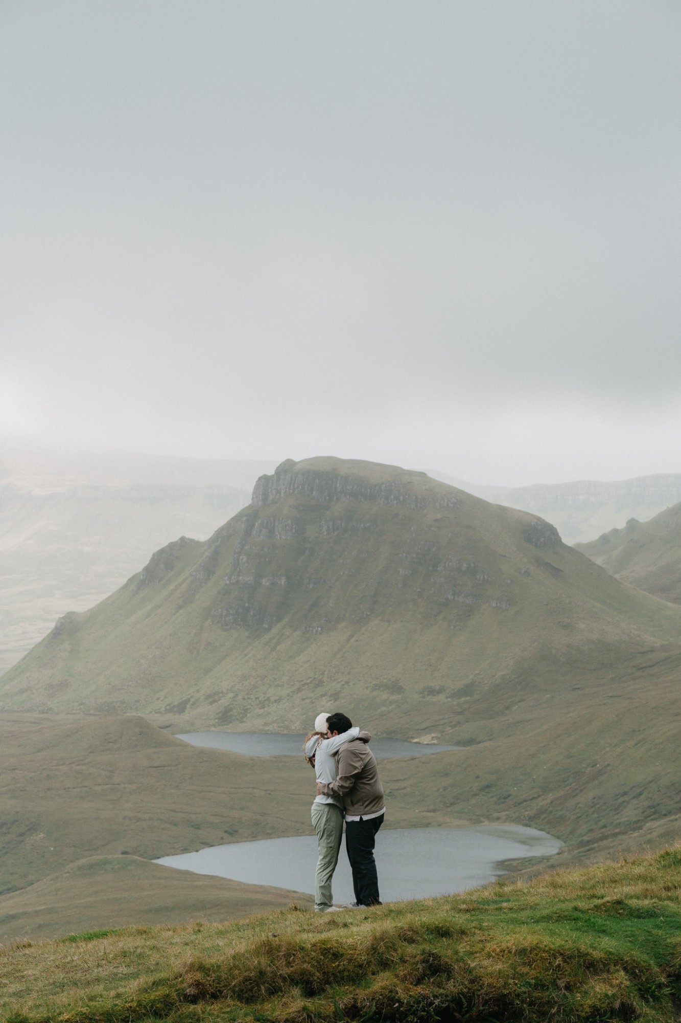 A couple embracing on a grassy ridge above two lochans at the Quiraing, Isle of Skye, a misty hill behind them