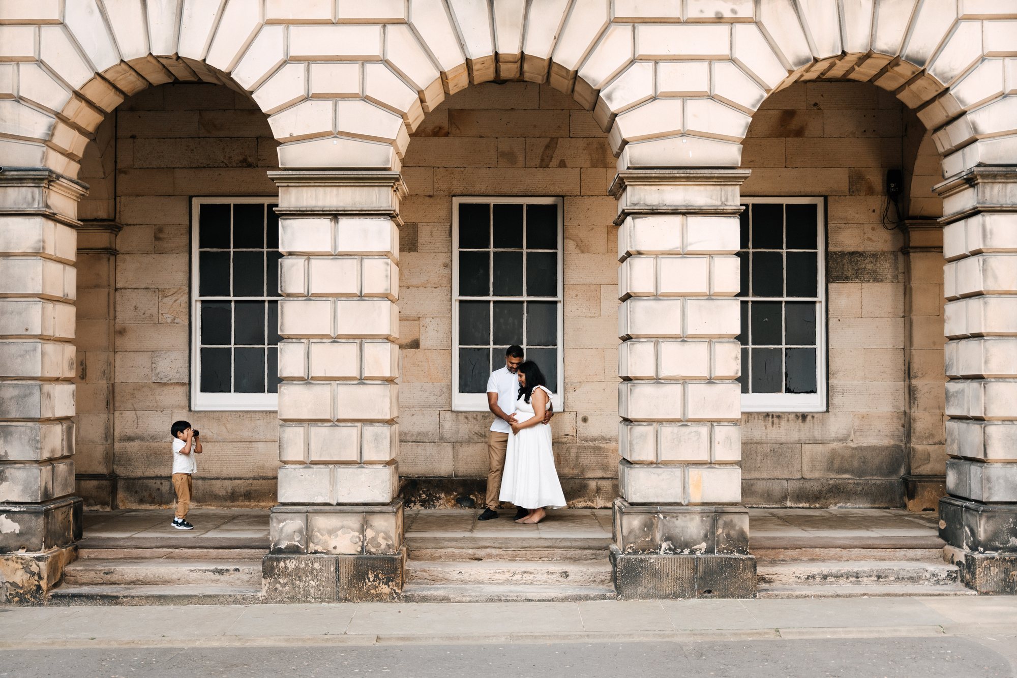 A couple expecting a baby under stone arches in Edinburgh while their young son takes their photograph