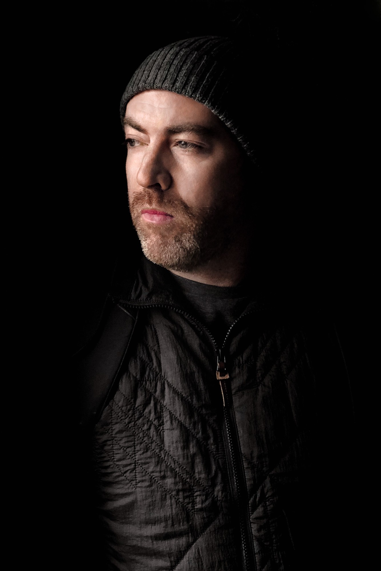 A bearded man in a knitted beanie and quilted jacket, looking off to the side in low light