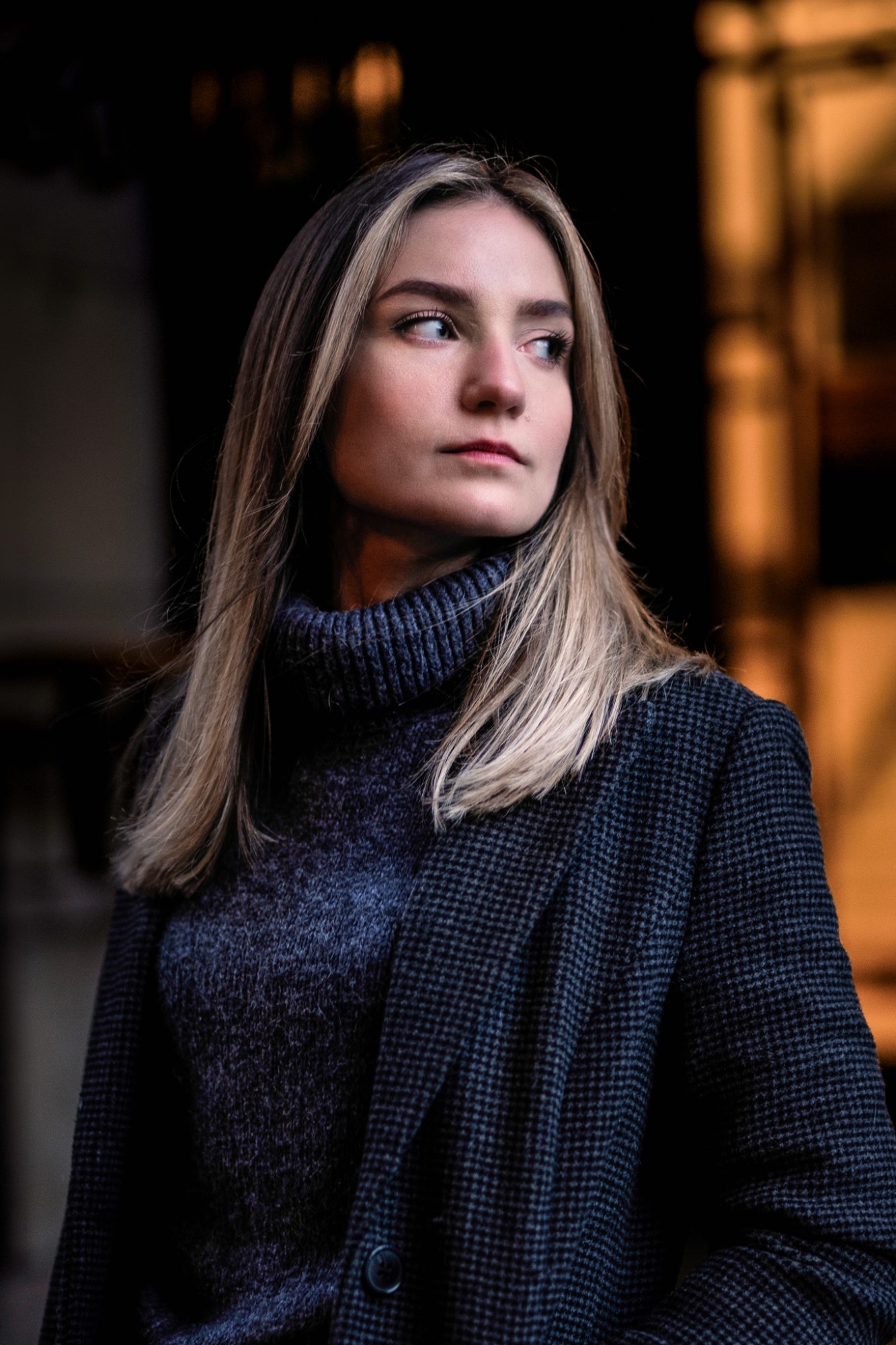 A woman in a grey roll-neck and checked coat, looking over her shoulder
