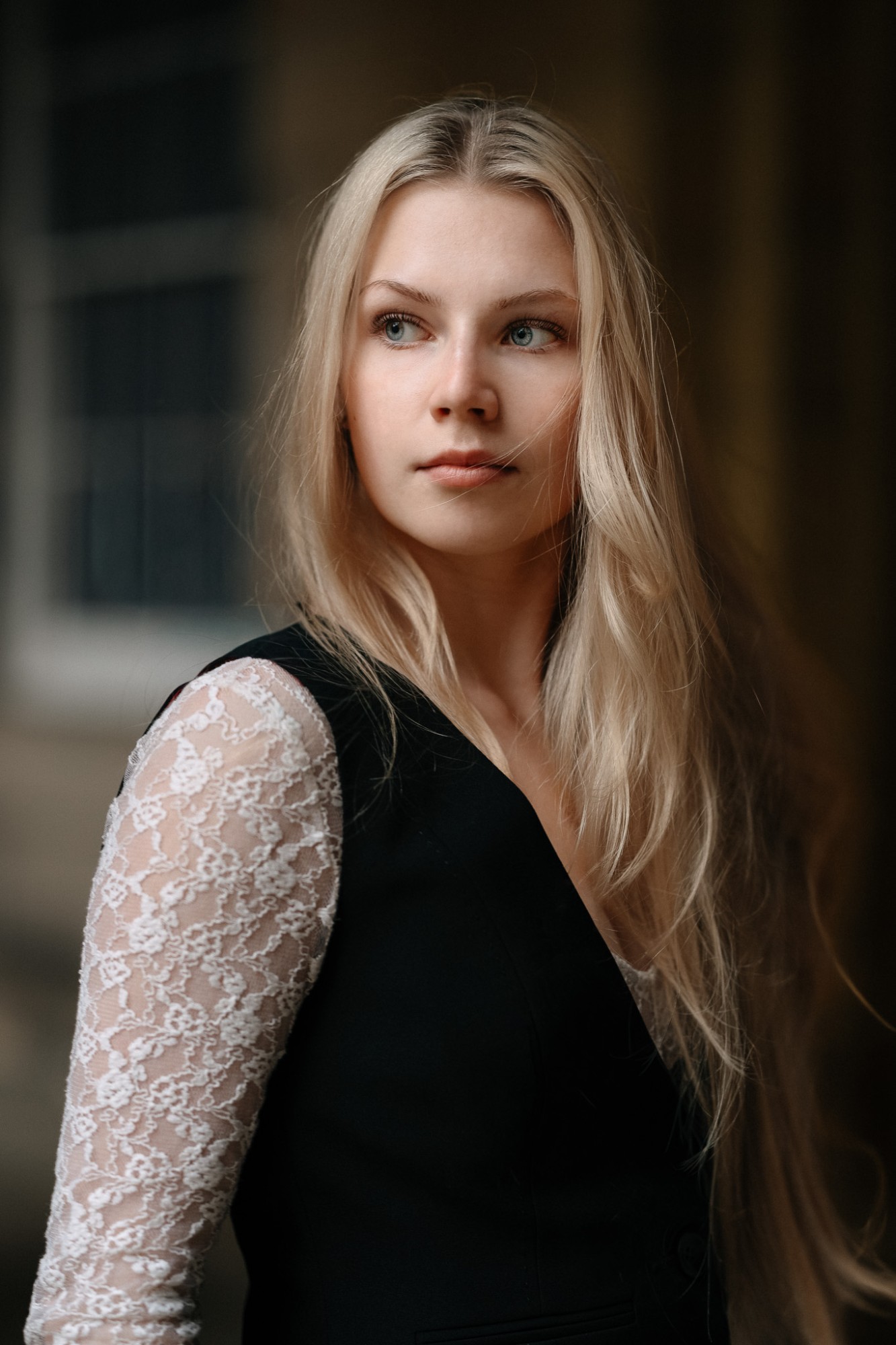 A woman with long blonde hair in a black waistcoat and white lace sleeves, looking off to the side