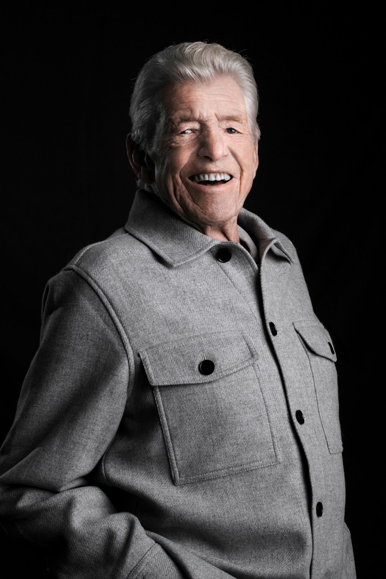 An older man with swept-back white hair laughing, in a grey overshirt against black