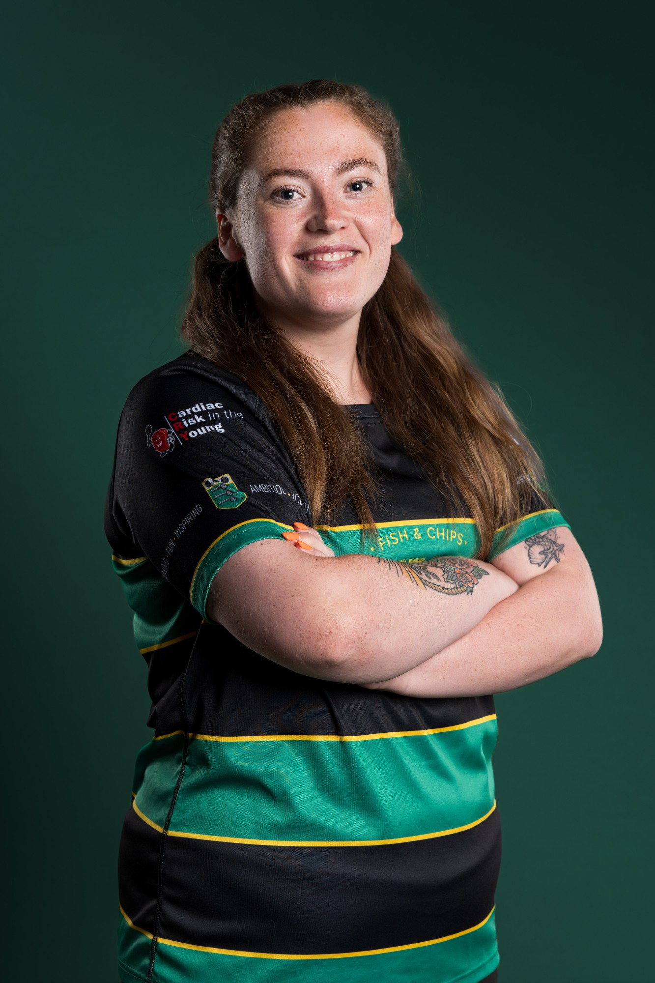 A smiling woman in a Bishopton RFC shirt, arms folded, against a dark green backdrop
