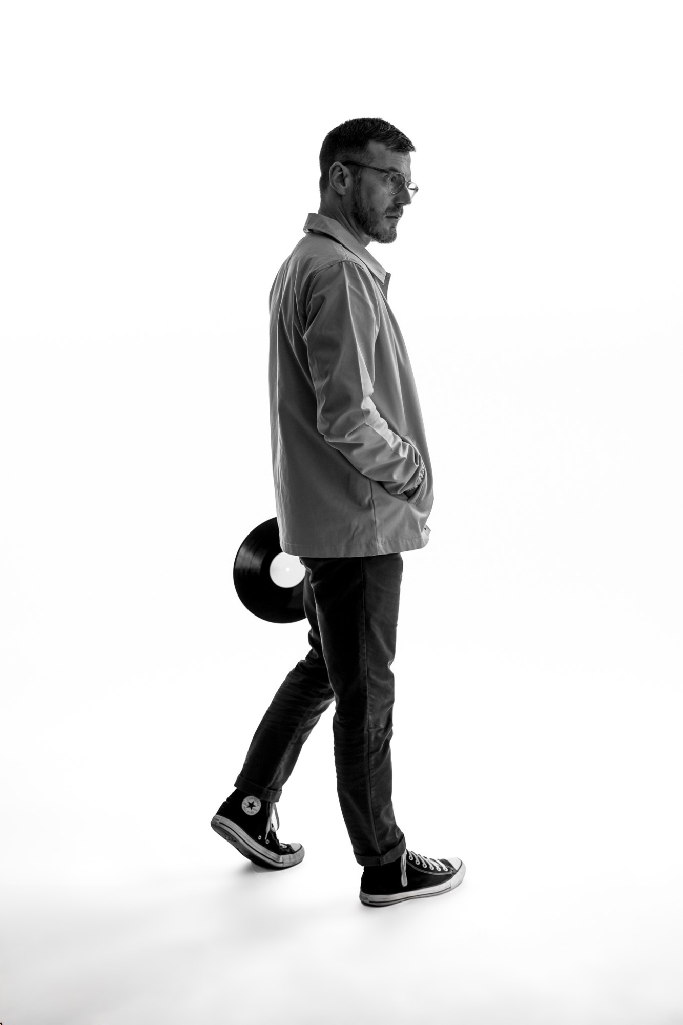 A man in glasses walking against a white background, a vinyl record in his hand, in black and white